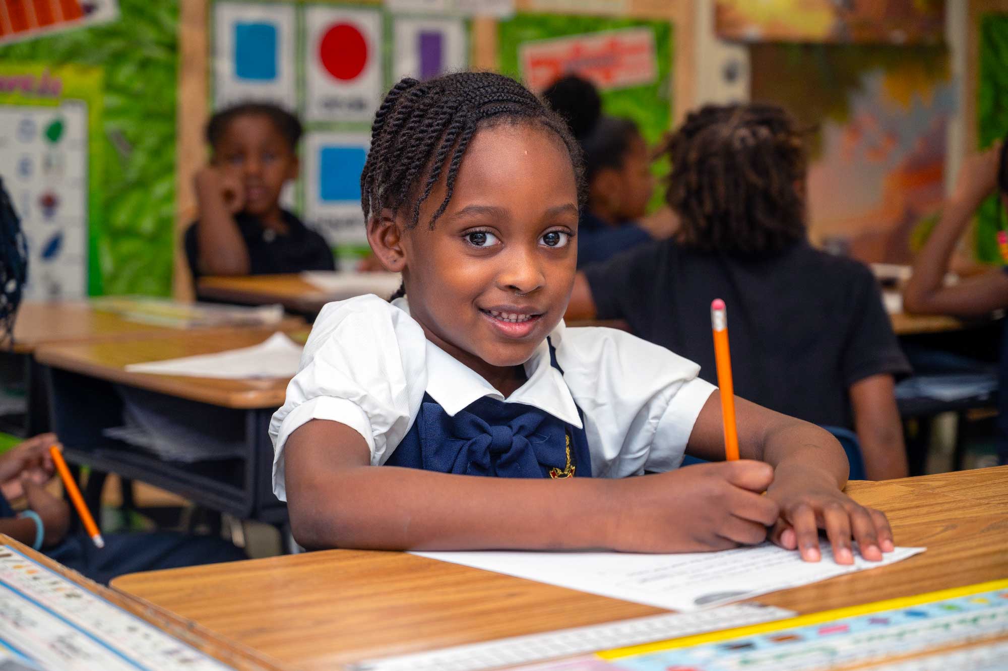 Smiling student writing with a pencil at a classroom desk.