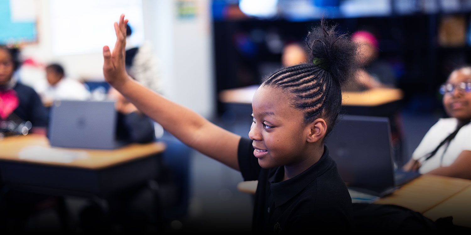 Smiling student with raised hand in a classroom.